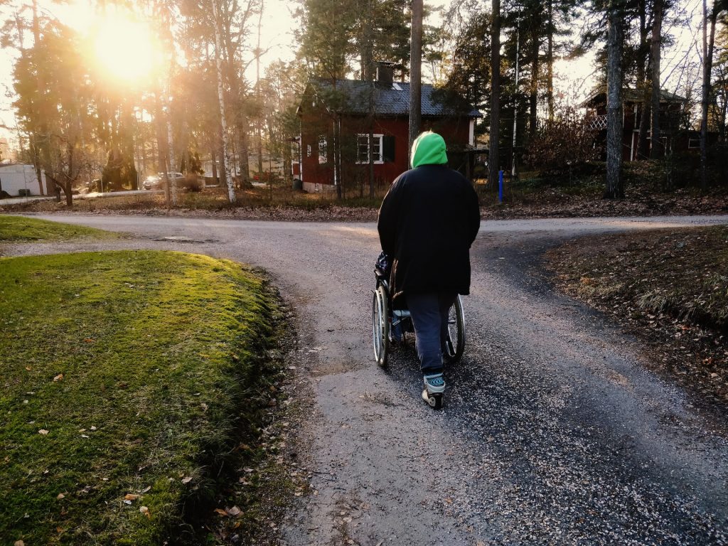 Takaapäin kuvattu henkilö työntää pyörätuolissa olevaa toista henkilöä pitkin hiekkatietä. Maisemassa näkyy risteys, havupuita ja punaisia puurakennuksia. Auringon valo siivilöityy puiden läpi.